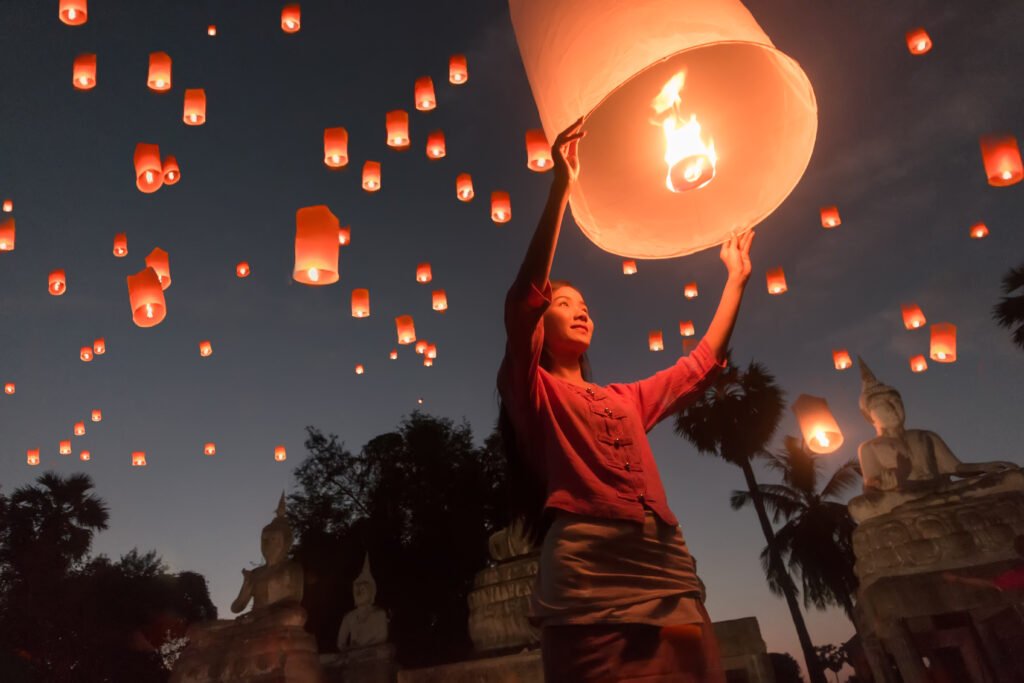 Person releasing a glowing sky lantern into the night sky during the Yi Peng Lantern Festival in Chiang Mai, Thailand, with dozens of lanterns floating overhead.