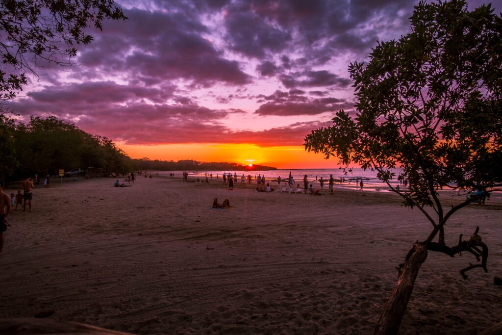 Families relaxing on Tamarindo Beach at sunset under a colorful sky.
