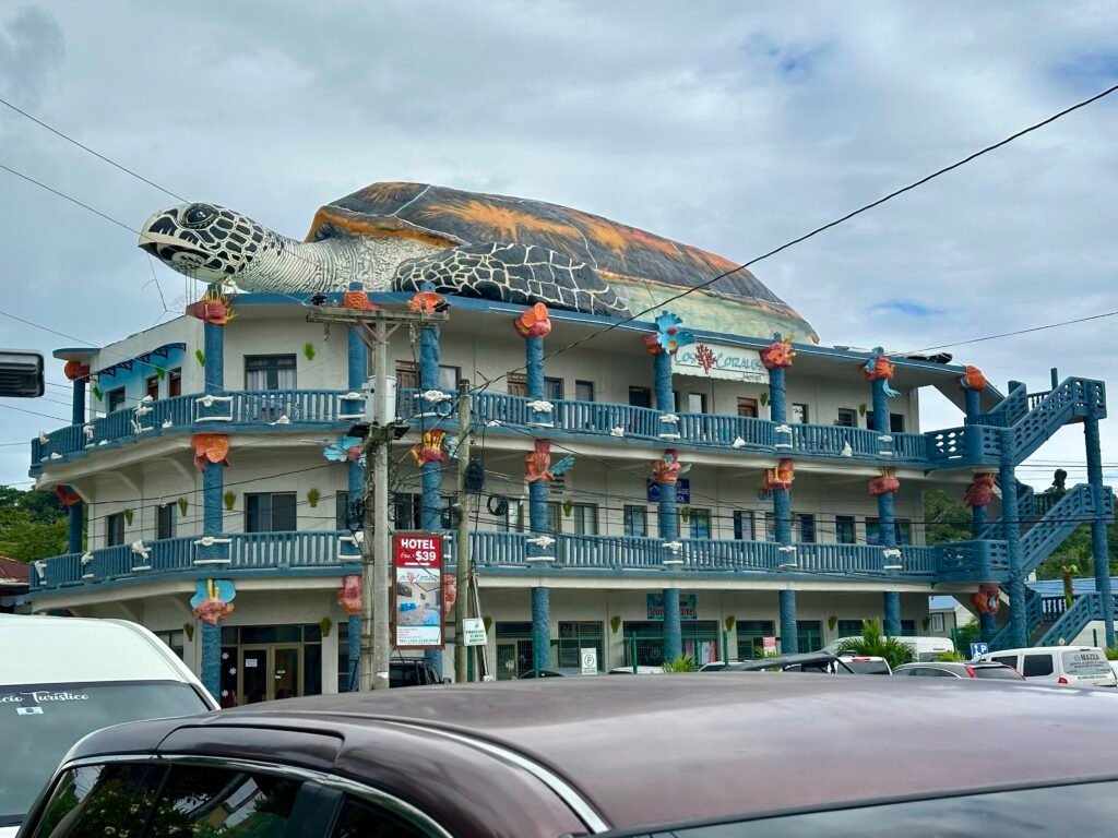 Colorful three-story building in Roatán topped with a giant turtle sculpture, seen from a car on the main road.