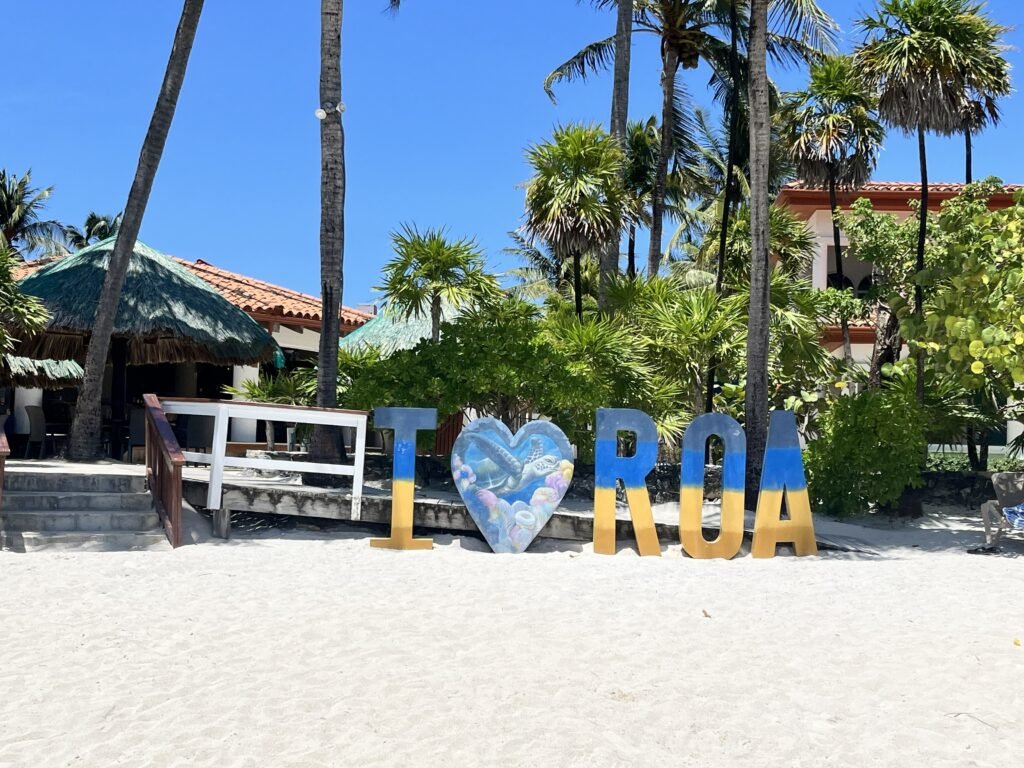 Colorful “I love Roa” sign on a sandy beach in Roatán, with palm trees and beachfront villas in the background.