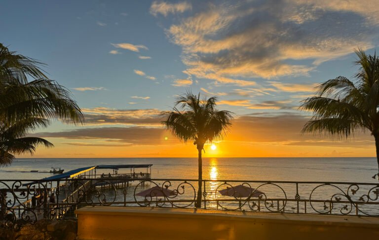 Sunset over West Bay Beach in Roatán, seen from a terrace with palm trees and a pier.