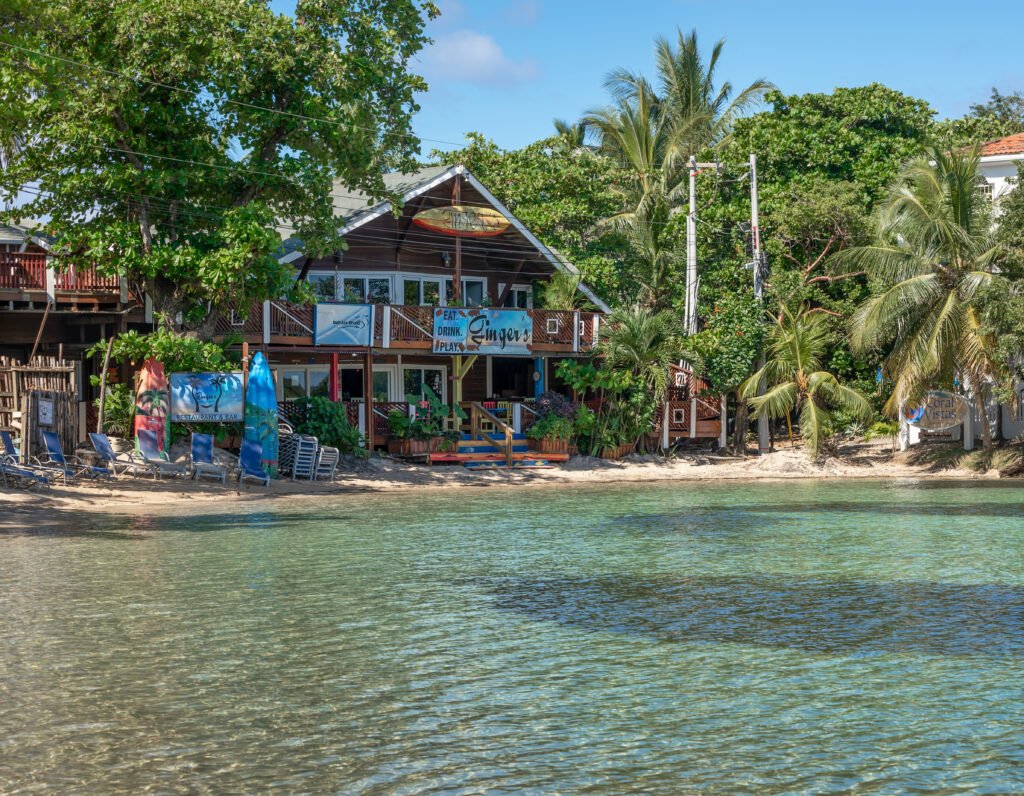 Ginger’s Caribbean Grill on the waterfront in West End, Roatán, with beach chairs and calm turquoise water in front.