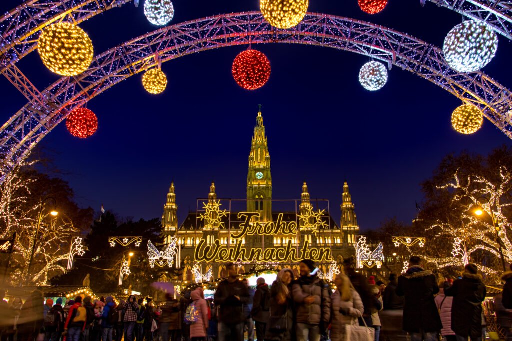 Visitors gather beneath illuminated ornaments and a Frohe Weihnachten sign at Vienna’s Rathausplatz Christmas Market.