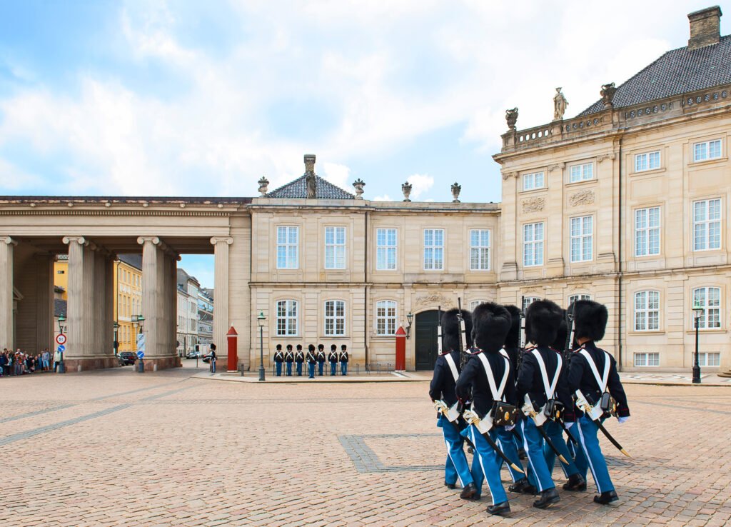 Royal guards in dark jackets, blue trousers, and tall black hats marching across the cobblestone square at Amalienborg Palace in Copenhagen.