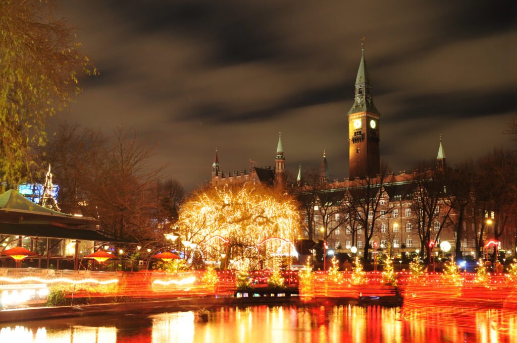 Golden Christmas lights and red light trails reflected in water beneath a clock tower in Copenhagen.