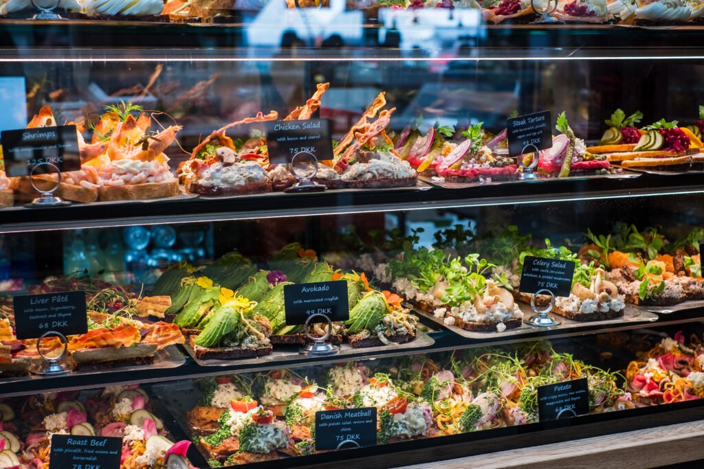 A glass display filled with Danish open-faced sandwiches topped with shrimp, avocado, meats, vegetables, and fresh herbs.