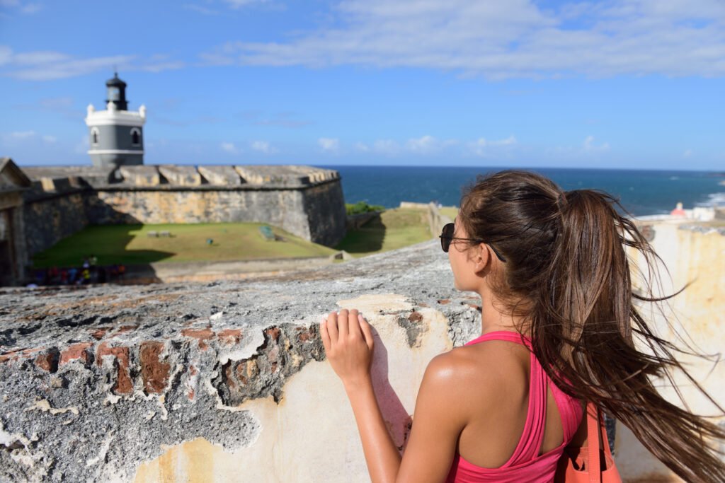 Visitor looking over a weathered stone wall toward El Morro’s lighthouse, fort grounds, and ocean.