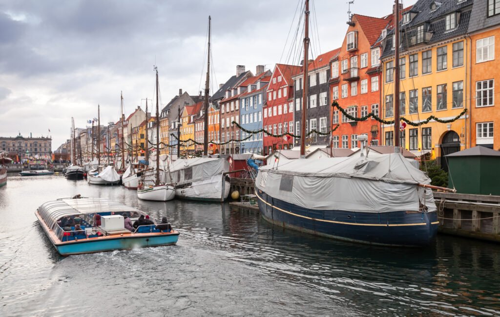 A sightseeing boat passing moored vessels and colorful waterfront buildings decorated with holiday garlands in Nyhavn, Copenhagen.