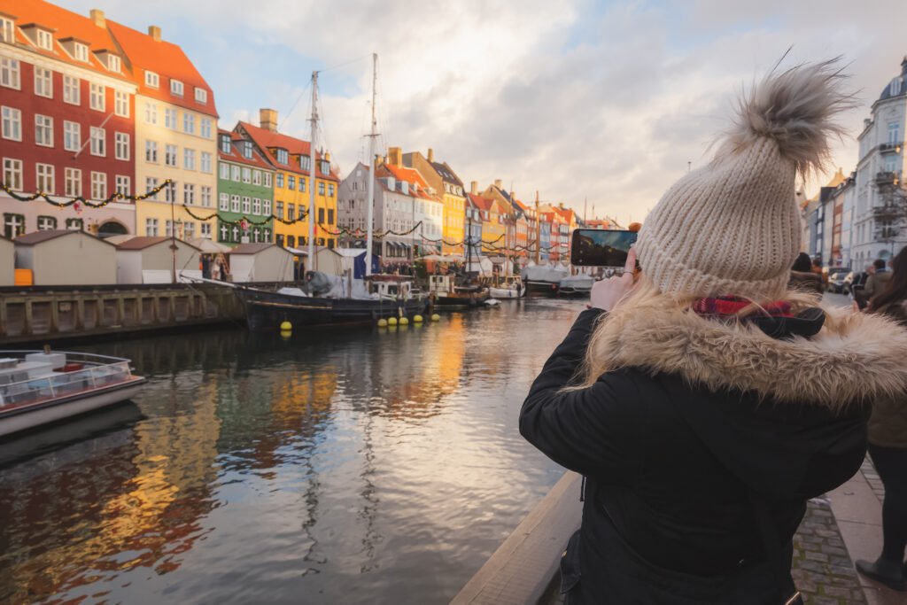 A visitor in a winter hat photographing Nyhavn’s colorful waterfront buildings, moored boats, and holiday garlands in Copenhagen.