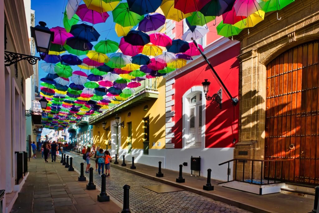Colorful umbrellas hang above a cobblestone street lined with bright buildings as visitors stroll beneath them.