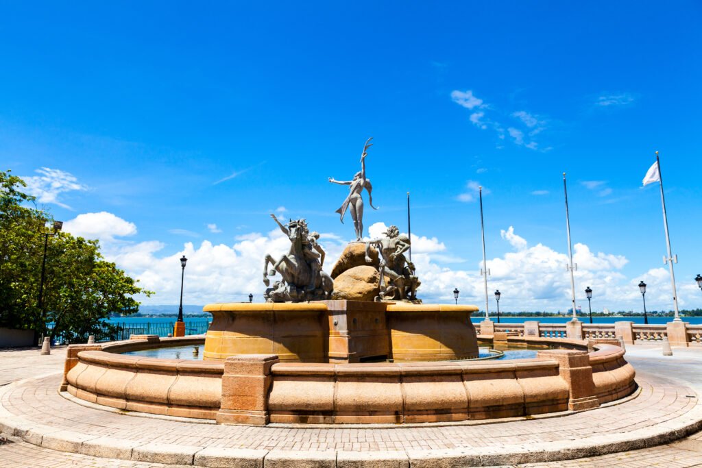 Sculptural fountain in a circular stone basin beside the waterfront, beneath a bright blue sky.