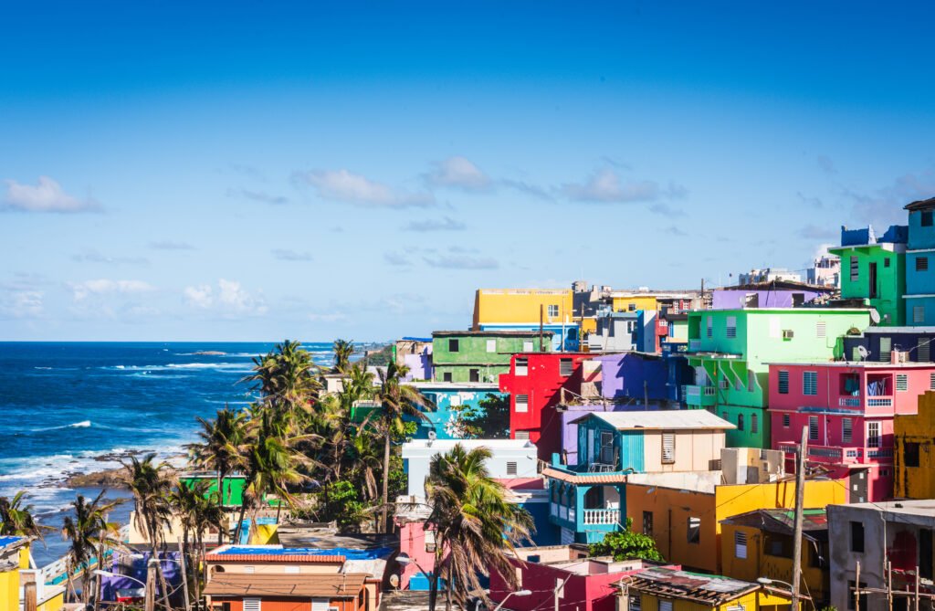 Colorful coastal buildings and palm trees overlook the blue ocean beneath a bright sky.
