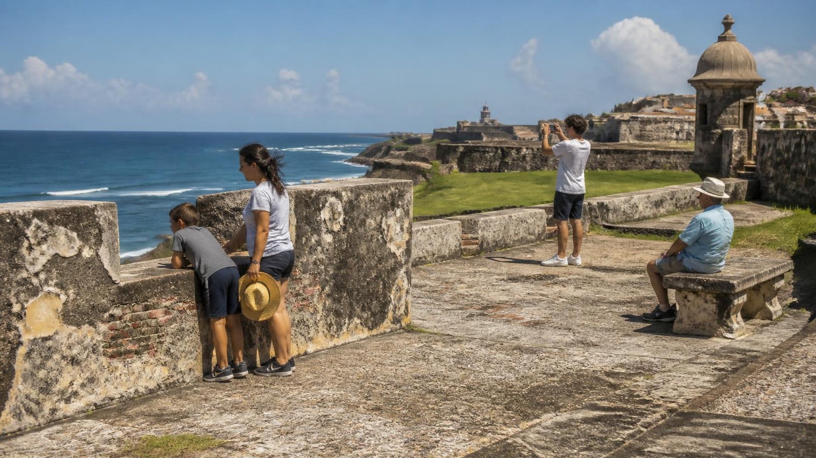 Multigenerational family exploring an oceanfront fort, with a child looking through a stone wall, a teen taking photos, and an older adult resting.