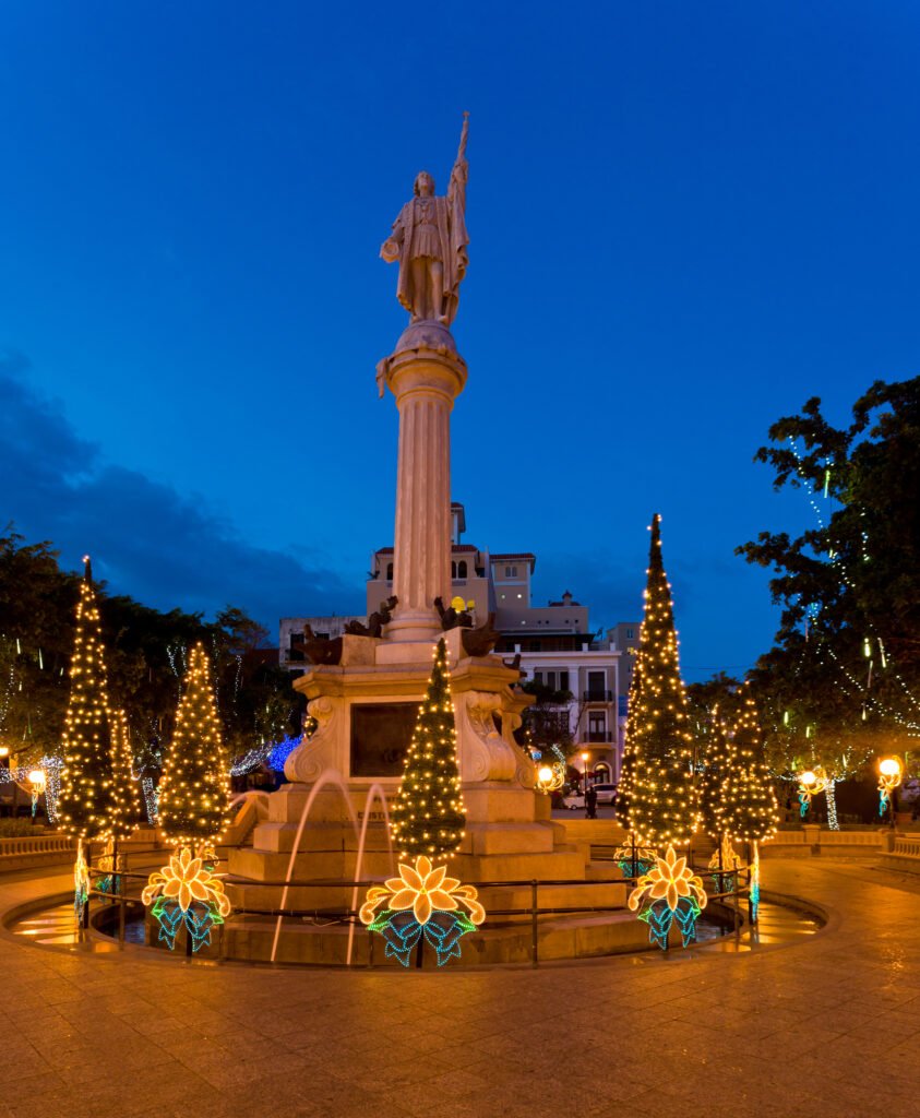Christmas lights and illuminated floral decorations surrounding a historic monument in a plaza at dusk.