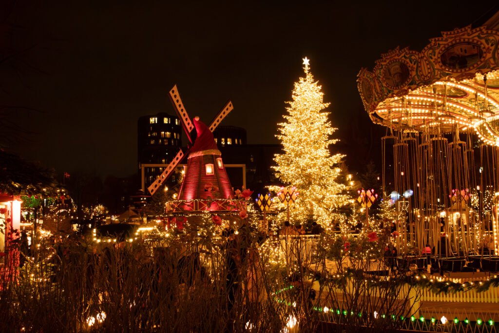 An illuminated Christmas tree beside a red windmill and ornate swing ride at Tivoli Gardens in Copenhagen at night.